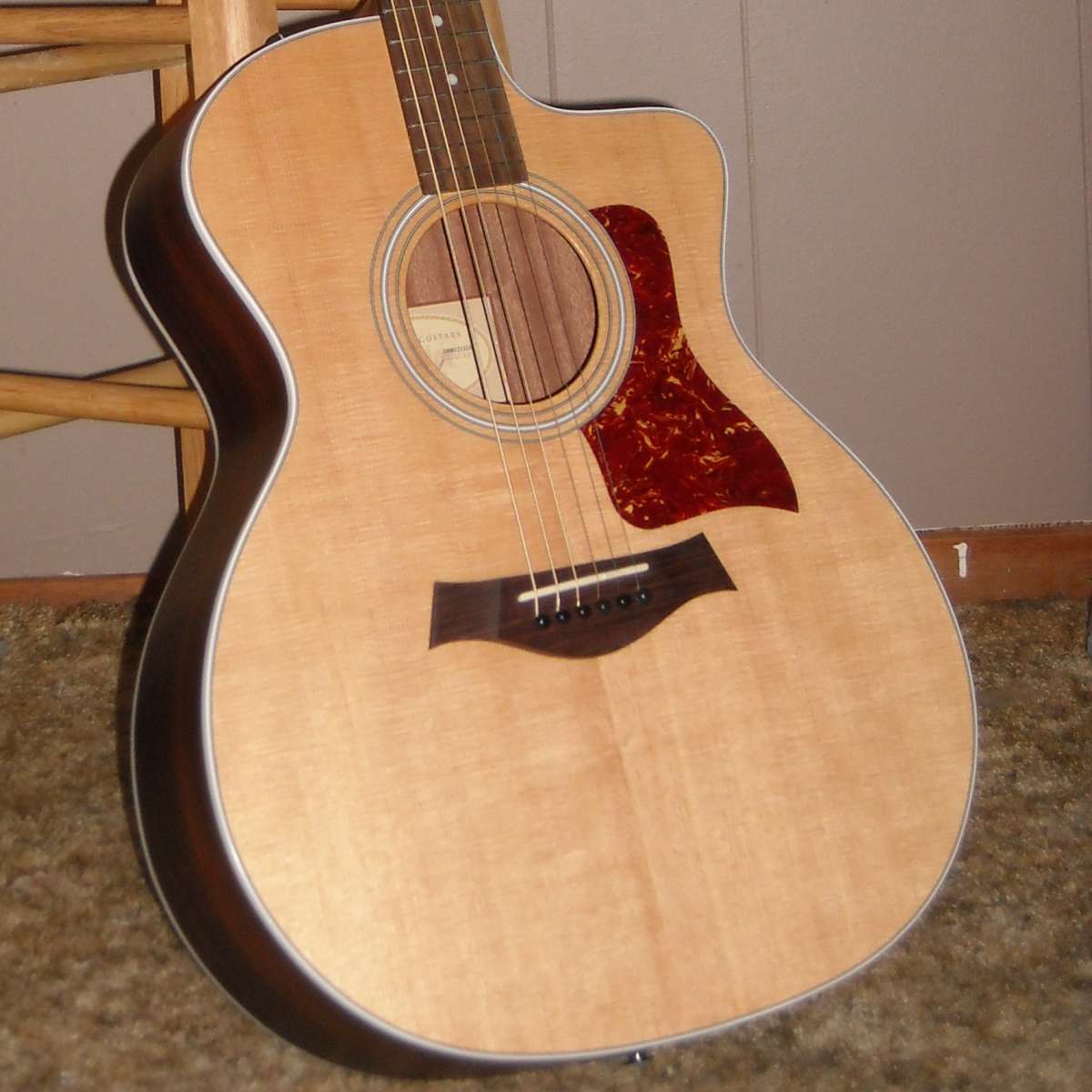 An image of a Taylor 214-CE Grand Concert acoustic guitar leaning against a stool