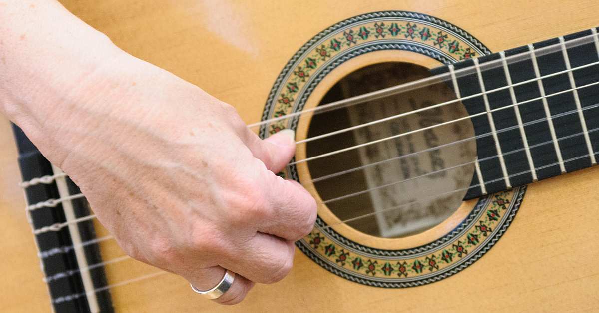 A thumb plucking the sixth string of the classical guitar and resting against the fifth string